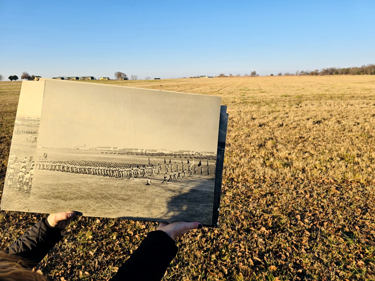 A hand holds a vintage panoramic photograph of massed troops on the Camp Gruber parade ground against the same empty field today